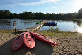 A sunny lakeside scene with three red kayaks resting on sandy ground near the shore. A wooden dock extends into the calm water, where several colorful paddle boats are moored. The lake is encircled by lush green trees, and the skies are clear with a few wispy clouds.