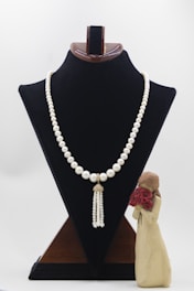 A pearl necklace with a tassel pendant is displayed on a black velvet stand. Beside the stand, there is a small figurine of a person holding a bouquet of red flowers.