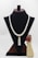 A pearl necklace with a tassel pendant is displayed on a black velvet stand. Beside the stand, there is a small figurine of a person holding a bouquet of red flowers.