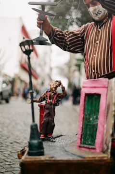A marionette puppet is being operated by a person wearing a striped shirt. The puppet is positioned on a small stage with a lamp post and a red mailbox as part of the scenery. In the background, there is an out-of-focus cobblestone street lined with buildings.
