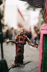 A marionette puppet is suspended by strings, featuring a detailed wooden face and hands. It is dressed in a brown outfit with red suspenders, standing on a textured surface. The puppet holds a small bottle in one hand. The background is blurred, depicting an outdoor scene in a city with hints of people and buildings.