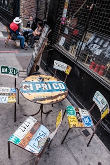 A unique outdoor cafe setting with a round table and chairs made from recycled license plates. Three people are sitting at the table, one wearing a white hat and red scarf. The background features a brick wall and a caged window displaying artwork and various objects.