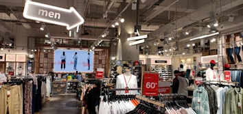 The interior of a retail clothing store featuring rows of clothing racks. A large digital screen at the back displays images of models wearing outfits. Hanging signs indicate sections for footwear and men's clothing. Bright overhead lighting and modern industrial decor contribute to the store's ambiance. Sales signs promote discounts, including a notable 60% off message.