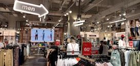 The interior of a retail clothing store featuring rows of clothing racks. A large digital screen at the back displays images of models wearing outfits. Hanging signs indicate sections for footwear and men's clothing. Bright overhead lighting and modern industrial decor contribute to the store's ambiance. Sales signs promote discounts, including a notable 60% off message.