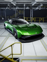A sleek green sports car with aerodynamic features is parked in an industrial setting. The car has a low profile and glossy exterior, and it is positioned inside a spacious facility with overhead lighting and yellow railings.