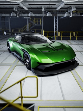 A sleek green sports car with aerodynamic features is parked in an industrial setting. The car has a low profile and glossy exterior, and it is positioned inside a spacious facility with overhead lighting and yellow railings.