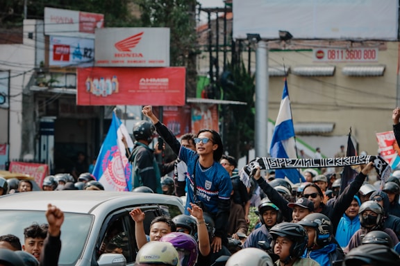 A large group of people, mostly wearing helmets, are gathered, with some waving flags and one person standing on a vehicle holding a banner. The setting appears to be a street in an urban area, with signs and buildings visible in the background.