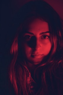 A close-up portrait of a person with long hair bathed in deep red lighting, creating dramatic shadows and highlights on the facial features.