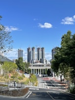 A cityscape with tall skyscrapers under construction in the background, surrounded by cranes. In the foreground, there is a modern plaza with greenery and pathways. A clear blue sky with a few clouds is visible, enhancing the urban landscape.
