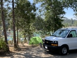 A campsite set in a forested area, featuring a green tent near a body of water with boats visible in the background. The scene includes a white van parked nearby and is surrounded by tall trees and natural greenery.