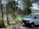 A campsite set in a forested area, featuring a green tent near a body of water with boats visible in the background. The scene includes a white van parked nearby and is surrounded by tall trees and natural greenery.