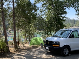 A campsite set in a forested area, featuring a green tent near a body of water with boats visible in the background. The scene includes a white van parked nearby and is surrounded by tall trees and natural greenery.