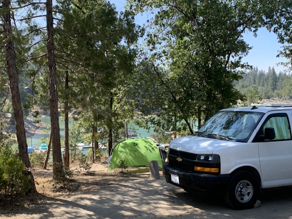 A campsite set in a forested area, featuring a green tent near a body of water with boats visible in the background. The scene includes a white van parked nearby and is surrounded by tall trees and natural greenery.