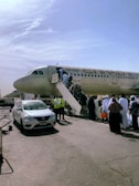 A passenger aircraft with people boarding via a stairway on an airport tarmac. The plane has Etihad Airways branding visible and there are ground crew members wearing high-visibility vests nearby. Several people, some dressed in traditional clothing, are lined up to get on the aircraft. A white car with an airport license plate is parked next to the scene.