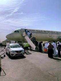 A passenger aircraft with people boarding via a stairway on an airport tarmac. The plane has Etihad Airways branding visible and there are ground crew members wearing high-visibility vests nearby. Several people, some dressed in traditional clothing, are lined up to get on the aircraft. A white car with an airport license plate is parked next to the scene.