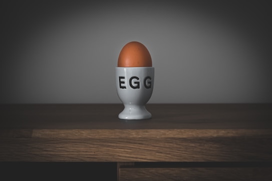 An egg rests upright in a white egg cup with the word 'EGG' printed on it in bold black letters. The scene is set on a wooden surface against a plain, dark background, creating a minimalist and slightly moody atmosphere.