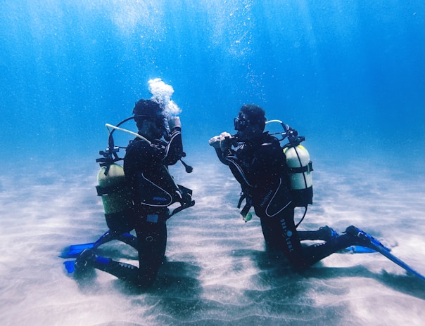 Two scuba divers are kneeling on the ocean floor facing each other, surrounded by a deep blue underwater environment. They are wearing full diving gear, including oxygen tanks, fins, and masks. Sunlight filters down through the water, creating patterns on the sandy bottom.