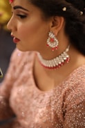 A woman is wearing elaborate jewelry including dangling earrings and a choker necklace adorned with red and silver elements. Her makeup is detailed, with carefully applied eyeliner and lipstick. She wears an embroidered pinkish outfit that shimmers, suggesting a formal or festive occasion. The close-up perspective emphasizes the elegance and intricacy of her attire and accessories.