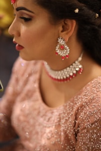 A woman is wearing elaborate jewelry including dangling earrings and a choker necklace adorned with red and silver elements. Her makeup is detailed, with carefully applied eyeliner and lipstick. She wears an embroidered pinkish outfit that shimmers, suggesting a formal or festive occasion. The close-up perspective emphasizes the elegance and intricacy of her attire and accessories.
