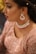 A woman is wearing elaborate jewelry including dangling earrings and a choker necklace adorned with red and silver elements. Her makeup is detailed, with carefully applied eyeliner and lipstick. She wears an embroidered pinkish outfit that shimmers, suggesting a formal or festive occasion. The close-up perspective emphasizes the elegance and intricacy of her attire and accessories.