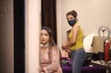 A woman dressed in an elegant pink outfit with intricate jewelry sits in a room, possibly preparing for a special event. Beside her, a person wearing a yellow tank top and jeans with a face mask is standing, suggesting assistance or preparation. Various beauty and grooming tools are visible on a table in the background, alongside a ring light, indicating a setting focused on beauty or fashion.