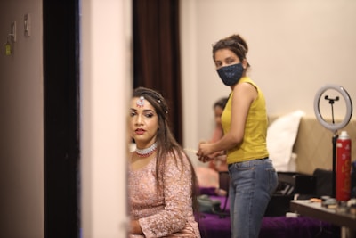 A woman dressed in an elegant pink outfit with intricate jewelry sits in a room, possibly preparing for a special event. Beside her, a person wearing a yellow tank top and jeans with a face mask is standing, suggesting assistance or preparation. Various beauty and grooming tools are visible on a table in the background, alongside a ring light, indicating a setting focused on beauty or fashion.