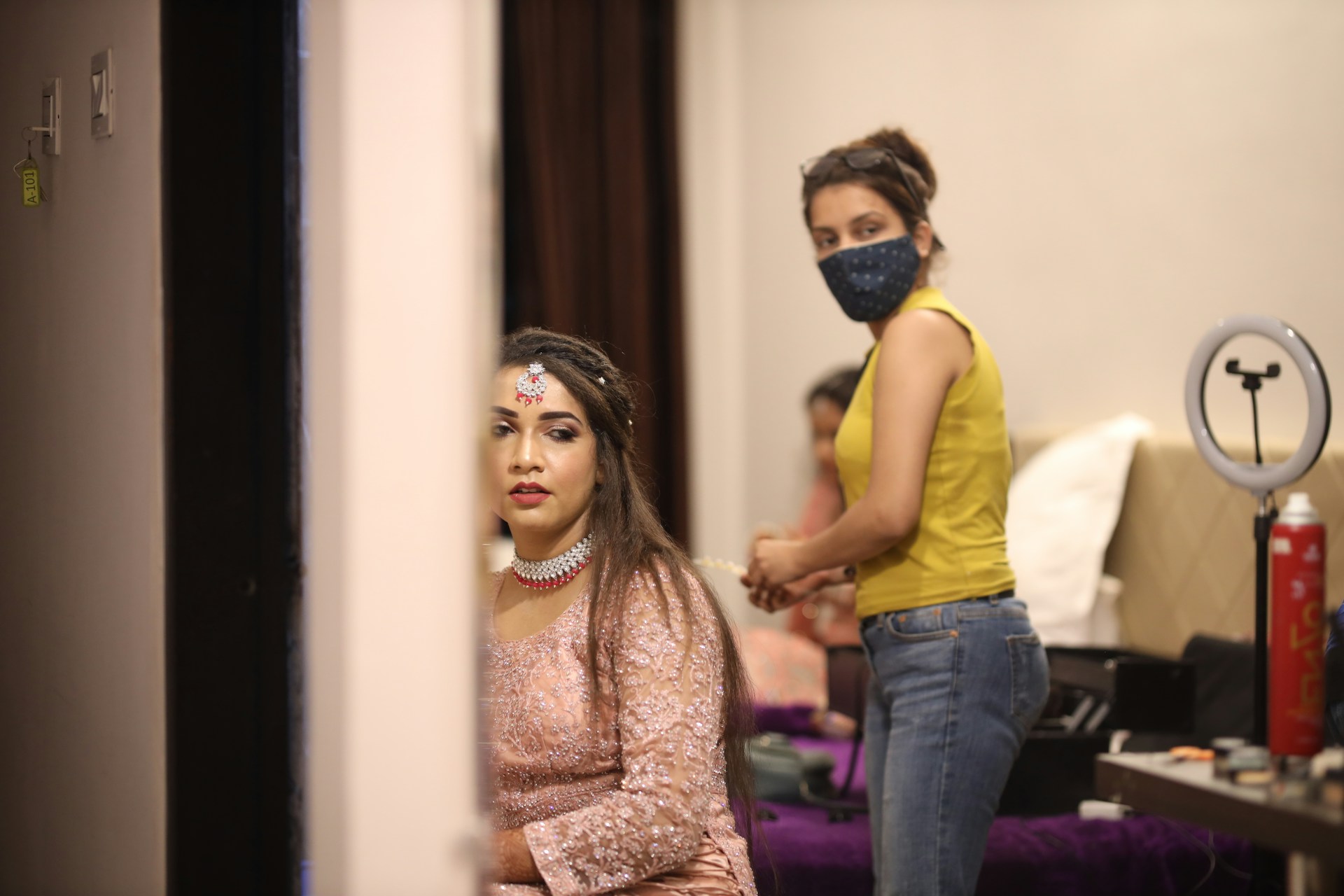 A woman dressed in an elegant pink outfit with intricate jewelry sits in a room, possibly preparing for a special event. Beside her, a person wearing a yellow tank top and jeans with a face mask is standing, suggesting assistance or preparation. Various beauty and grooming tools are visible on a table in the background, alongside a ring light, indicating a setting focused on beauty or fashion.