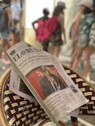 A newspaper is resting on a woven chair, prominently showing a front-page article with a photo of a man in a suit covering his face. The background is blurred, with several people walking, some carrying backpacks and wearing casual summer clothing.