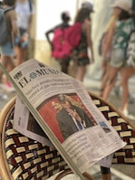 A newspaper is resting on a woven chair, prominently showing a front-page article with a photo of a man in a suit covering his face. The background is blurred, with several people walking, some carrying backpacks and wearing casual summer clothing.