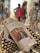 A newspaper is resting on a woven chair, prominently showing a front-page article with a photo of a man in a suit covering his face. The background is blurred, with several people walking, some carrying backpacks and wearing casual summer clothing.