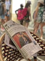 A newspaper is resting on a woven chair, prominently showing a front-page article with a photo of a man in a suit covering his face. The background is blurred, with several people walking, some carrying backpacks and wearing casual summer clothing.