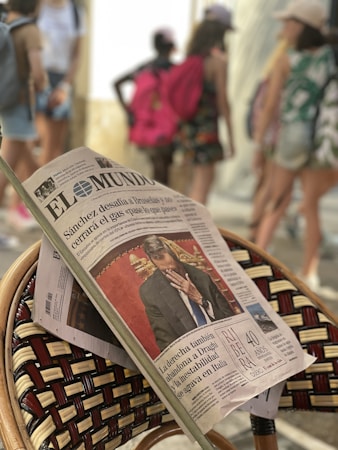A newspaper is resting on a woven chair, prominently showing a front-page article with a photo of a man in a suit covering his face. The background is blurred, with several people walking, some carrying backpacks and wearing casual summer clothing.