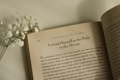 An open book lies on a flat surface with visible text on the page, featuring the heading 'Loving Oneself as the Path to the Divine.' Accompanied by a small sprig of white flowers resting on the left side of the book.