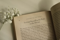 An open book lies on a flat surface with visible text on the page, featuring the heading 'Loving Oneself as the Path to the Divine.' Accompanied by a small sprig of white flowers resting on the left side of the book.