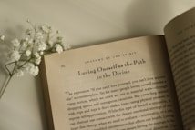 An open book lies on a flat surface with visible text on the page, featuring the heading 'Loving Oneself as the Path to the Divine.' Accompanied by a small sprig of white flowers resting on the left side of the book.