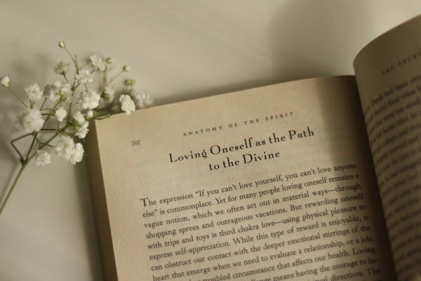 An open book lies on a flat surface with visible text on the page, featuring the heading 'Loving Oneself as the Path to the Divine.' Accompanied by a small sprig of white flowers resting on the left side of the book.