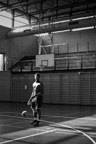 A black and white photograph of a shirtless young man standing in a gymnasium holding a basketball. Sunlight streams through large windows, casting geometric shadows on the wooden floor. The background includes a basketball hoop and bleachers.