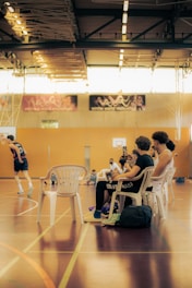 Inside a spacious gymnasium, several people sit on white plastic chairs observing a basketball game. One individual is using a smartphone to record or take pictures while players are in motion on the court. The environment is filled with natural light streaming through large windows, and the walls are decorated with banners.
