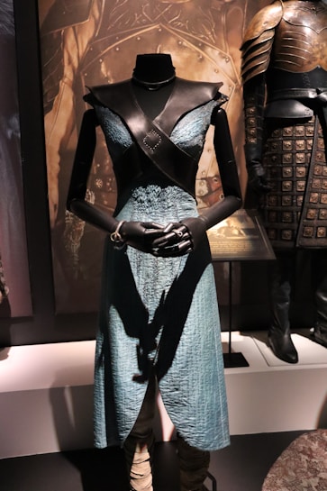 A black mannequin wears a medieval-style costume featuring a blue textured dress with a black leather bodice and black gloves. The background includes another piece of armor displayed nearby, emphasizing a historical or fantasy theme.