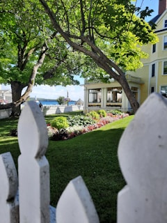 A picturesque garden scene featuring a well-maintained lawn with colorful flowerbeds. Large, leafy trees provide ample shade, and a charming yellow building with white windows stands in the background. A white picket fence is in the foreground, and a glimpse of the water and a blue sky can be seen beyond the garden.