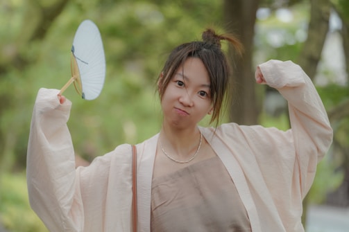 A person wearing a light, flowing kimono stands outdoors, playfully holding a fan in one hand. The background is blurred with lush green foliage, suggesting a garden setting.