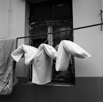 Bed sheets and pillow covers are hanging on a clothesline in front of an old, weathered window. The window has peeling paint and looks aged. The wall behind the clothesline is two-toned with a darker bottom half.