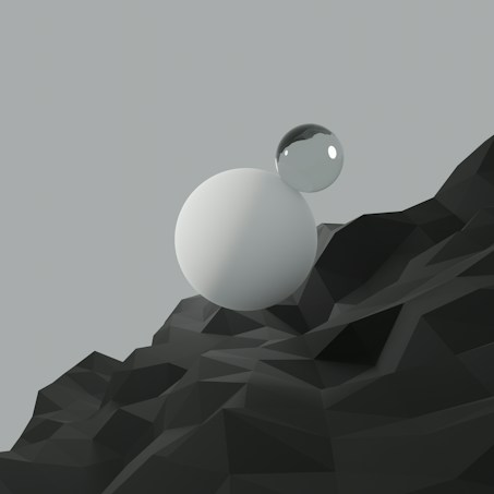 A minimalistic landscape featuring two spheres, one opaque and one transparent, resting on a low-poly styled, geometric surface with sharp edges. The background is a soft grey color, contributing to a serene and modern aesthetic.