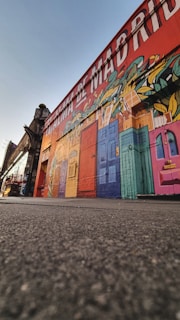 A vibrant mural decorates the side of a building, featuring various colorful doors and abstract designs. The wall is painted with images of leaves and patterns interwoven with the doors. Above, large text in white letters reads a phrase in Spanish. The scene is captured from a low-angle perspective, with the ground in the foreground and part of the street visible on the left.