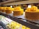 Multiple yellow frosted cakes with decorative flowers on top, displayed in a refrigerated showcase. Each cake is neatly arranged on a white paper base and surrounded by a variety of smaller desserts on lower shelves presented with reds, whites, and greens.