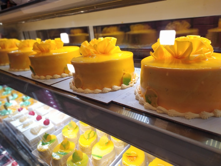Multiple yellow frosted cakes with decorative flowers on top, displayed in a refrigerated showcase. Each cake is neatly arranged on a white paper base and surrounded by a variety of smaller desserts on lower shelves presented with reds, whites, and greens.