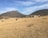 A vast, dry landscape with two brown cows grazing in an open field. In the background, there are two prominent hills against a blue sky with scattered clouds. A small, rustic building made of brick and metal stands near the hills, surrounded by sparse vegetation.
