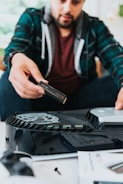A person in a plaid shirt is installing a storage drive into a gaming console, with focus on the electronics and assembly process.