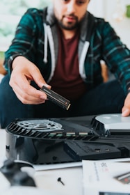 A person in a plaid shirt is installing a storage drive into a gaming console, with focus on the electronics and assembly process.