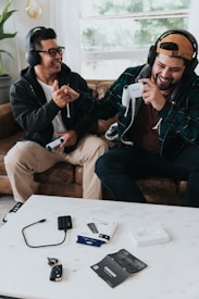 Two people are sitting on a couch wearing headphones and playing video games. They appear to be sharing a light-hearted moment and are engaged with the game controllers in their hands. On the table in front of them, there are electronic items including a Samsung box, a portable SSD, and a set of keys.